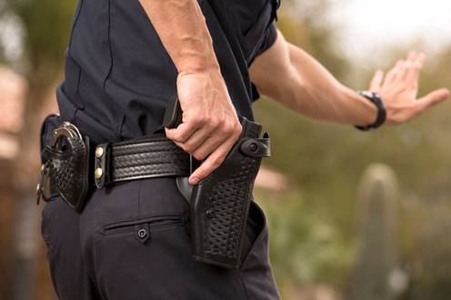 A police officer is shown from the rear, hand on their holstered pistol. Their other hand is raised in a stopping motion in an outdoor setting.